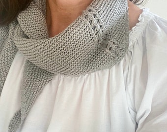 May include: A gray knitted scarf with a delicate lace pattern around the edges. The scarf is draped around a person's neck and is worn over a white shirt.
