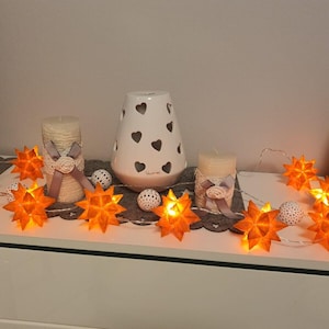 Könnte beinhalten: Eine Lichterkette aus orangefarbenen Papiersternen mit weißen Kerzen und einem weißen Keramikdiffusor mit Herz-Ausschnitten.