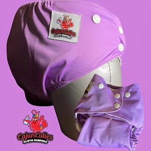 May include: A light purple cloth diaper with white snaps and a white label that says "Cajun Cuties Cloth Diapers" with a cartoon crawfish logo. The diaper is open, revealing the interior. The background is a solid purple.