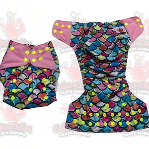 May include: Two cloth diapers with a colorful mermaid scale pattern. The diapers feature a pink trim and yellow snap closures. One diaper is open, and the other is closed. The text "Cajun Cuties Cloth Diapers" is visible.