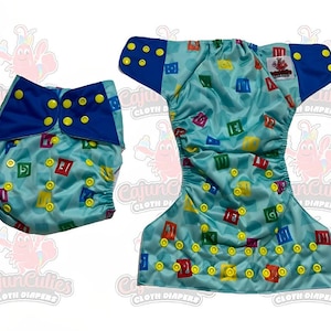 May include: Two cloth diapers. One is folded with a blue flap and yellow snaps. The other is open, showing a light blue background with colorful block letters and yellow snaps. The diapers are from Cajun Cuties.