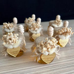 Pode incluir: Pequenos frascos de vidro cheios de mel dourado, cada um coberto com serapilheira, flores brancas secas e um pau de mel de madeira. Uma etiqueta de madeira em forma de coração gravada com o nome Safiya está presa na frente de cada frasco.