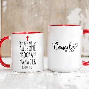 Puede incluir: Dos tazas de cerámica blancas con asas y bordes rojos. Una taza dice "THIS IS WHAT AN AWESOME PROGRAM MANAGER LOOKS LIKE" con una flecha. La otra taza dice "Camila EST 2026" con un corazón.