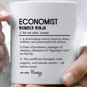 May include: White ceramic mug with the text "ECONOMIST NUMBER NINJA" and a definition. The definition includes phrases like "multitasking wizard" and "unofficial therapist." The mug is held by a person wearing gold rings.