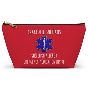 Può includere: Un astuccio rosso con cerniera con la scritta "CHARLOTTE WILLIAMS", un simbolo medico blu e le parole "SHELLFISH ALLERGY EMERGENCY MEDICATION INSIDE". L'astuccio ha una cerniera dorata.