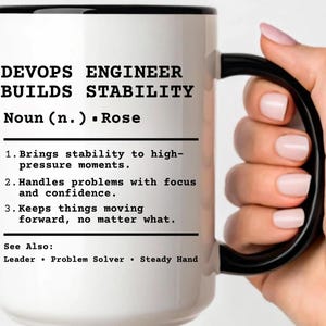 May include: White ceramic mug with a black handle and rim. The mug features the text "DEVOPS ENGINEER BUILDS STABILITY" and a definition. The mug is held by a person.