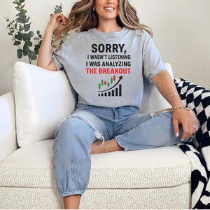 May include: A light gray t-shirt with the text "SORRY, I WASN'T LISTENING I WAS ANALYZING THE BREAKOUT" in black and red. The shirt features a graphic of a stock chart with an upward arrow. The person is wearing blue jeans.