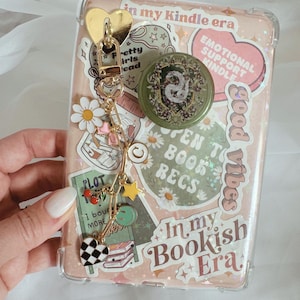 May include: A clear phone case decorated with stickers and a gold charm. Stickers include daisies, hearts, and text that reads "In my Kindle era," "Emotional Support Kindle," and "In my Bookish Era." The charm features a gold heart and various dangling charms.