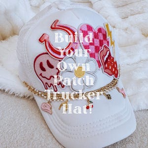 May include: White trucker hat with a mesh back and a curved brim. The hat is decorated with patches including a horseshoe, a skull, a heart, and a flower. The text "Build Your Own Patch Trucker Hat!" is displayed on the front. A gold chain with charms hangs from the brim.