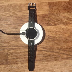 Op de afbeelding: Een zwarte smartwatch met een geperforeerde siliconen band rust op een ronde witte draadloze oplaadpad. Het horloge heeft een rond, donker scherm en een zilverkleurige gesp. Een zwarte oplaadkabel steekt uit de zijkant van de witte basis.