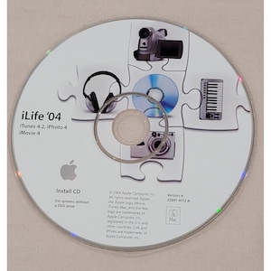 May include: A silver and white iLife '04 install CD. The disc features a puzzle design with images of a camera, headphones, a keyboard, and a camcorder. The Apple logo is visible, along with text indicating iTunes, iPhoto, and iMovie software.