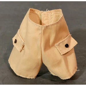 May include: Tan-colored doll shorts with two front pockets and black buttons. The shorts have a zipper and are made of a light fabric. The stitching is visible around the pockets and waistband. These shorts are designed for a doll.