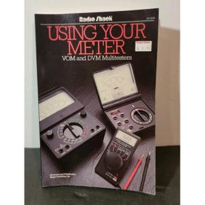 Puede incluir: Un libro vintage de Radio Shack titulado "Using Your Meter" con el subtítulo "VOM and DVM Multitesters". La portada presenta imágenes de multímetros. La portada del libro es negra con texto rojo y blanco.