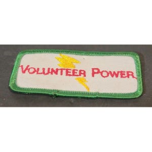 Könnte beinhalten: Gestickter Aufnäher mit den Worten "VOLUNTEER POWER" in roten Buchstaben auf weißem Grund. Der Aufnäher hat einen grünen Rand und gelbe Blitzdesigns. Ein rechteckiger Aufnäher.