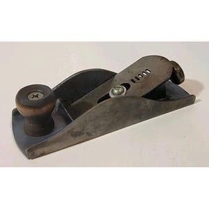 Puede incluir: Un cepillo de mano pequeño y antiguo, con un cuerpo gris oscuro y un pomo de madera. La cuchilla es visible y el cepillo parece ser de metal. Esta herramienta de carpintería tiene un aspecto clásico y usado, ideal para trabajar la madera.