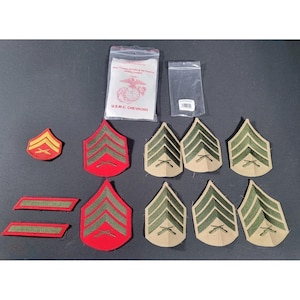May include: A collection of U.S.M.C. chevrons, including red and khaki-colored patches with green stripes and crossed rifles. Some chevrons are packaged in clear plastic bags. One bag has text that reads "U.S.M.C. CHEVRONS".