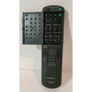 Könnte beinhalten: Eine schwarze, programmierbare Sony-Fernbedienung mit aufklappbarer Abdeckung. Die Fernbedienung verfügt über zahlreiche Tasten für TV- und VCR-Funktionen, darunter Ein/Aus, Lautstärke, Kanal und Menü. Der Text "SONY PROGRAMMABLE COMMANDER RM-Y704A" ist sichtbar.