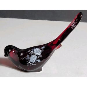 May include: A deep red glass bird figurine featuring hand-painted white floral details on its side and tail. The translucent ruby-coloured glass sculpture is shaped like a bird with a long, pointed tail and a smooth, curved body profile.
