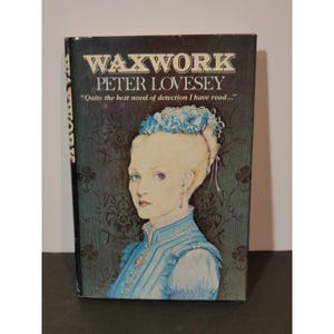 May include: A book cover for "Waxwork" by Peter Lovesey. The cover art depicts a woman in a blue dress and a floral headpiece. The title and author's name are in white, with a quote from a review below.