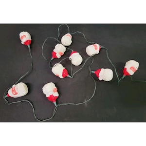 Könnte beinhalten: Eine Lichterkette mit zehn geformten Kunststoff-Weihnachtsmannköpfen. Jede Weihnachtsmannfigur ist weiß mit einer roten Mütze und bemalten Gesichtszügen. Die Figuren sind durch ein dünnes grünes Stromkabel für dekorative Zwecke verbunden.
