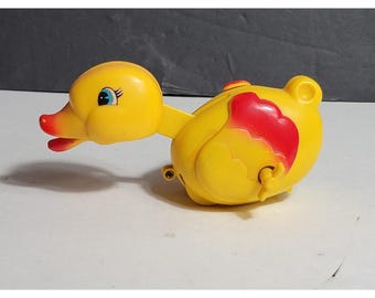 Peut inclure: Un jouet canard mécanique en plastique jaune avec un bec rouge et des ailes rouges. Le canard présente de grands yeux bleus avec des cils noirs et une petite clé de remontage dépassant du côté de son corps arrondi pour le mouvement.