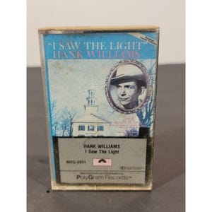 May include: Vintage cassette tape for Hank Williams' "I Saw The Light." The cassette case has a blue and white cover with the album title and artist's name. A portrait of Hank Williams is featured, along with a church illustration. The tape is from PolyGram Records.