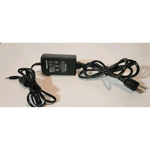 Puede incluir: Un adaptador de corriente Polaroid negro con un cable negro y un enchufe de dos clavijas. El adaptador tiene forma rectangular y está conectado a un enchufe más pequeño a la izquierda. El cable está enrollado.