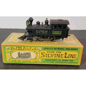 Puede incluir: Un modelo de tren negro con el texto "BA LTIMO RE AND OH 10" en el lateral, sobre una caja amarilla y verde. La caja dice "The B&O Teakettle" y "Quality HO Model Railroads From The Silvine Line".