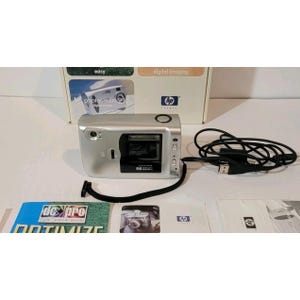 Puede incluir: Una cámara digital HP Photosmart plateada con un área de lente negra, un botón de obturador circular y una correa de muñeca negra. La cámara viene con un cable USB negro, una caja con la etiqueta easy digital imaging y manuales impresos.