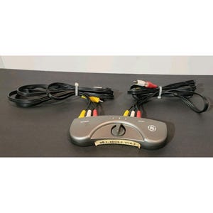 Puede incluir: Un selector de vídeo GE gris con múltiples puertos de entrada y salida RCA. El dispositivo tiene un dial y está conectado a cables negros con conectores rojos, blancos y amarillos. El texto en el dispositivo dice "Wii-1, XBOX 2, VCR 3."