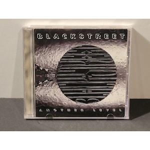 May include: A compact disc case featuring the album "Another Level" by Blackstreet. The cover art displays the band's name in bold white letters above a black and white circular design. The album title is printed in white at the bottom.