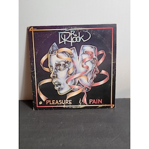 Puede incluir: Una portada de álbum de vinilo cuadrada para Dr. Hook titulada Pleasure & Pain. La portada presenta un fondo morado oscuro con una ilustración central de dos rostros estilizados de aspecto metálico entrelazados con cintas rosas y el texto.