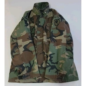 May include: Camouflage pattern jacket in shades of green, brown, and black. The jacket features a high collar, button and zipper closure, and a patch with the letters "U.S." and a crest. The jacket is made of a durable fabric.