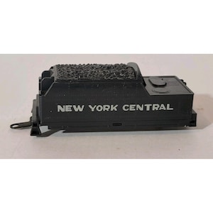 May include: A black model train tender with the words "NEW YORK CENTRAL" printed in white. The top of the tender has a textured surface, and the model has several detailed features.