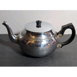 May include: A polished silver-tone metal teapot featuring a rounded body with a subtle hammered texture, a beaded decorative band around the shoulder, a black handle, and a matching black knob on the removable lid. The spout is curved and tapered.