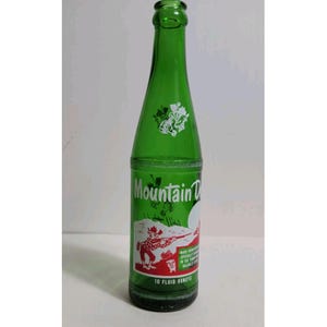 以下が含まれることがあります： 細い首を持つヴィンテージの緑色のガラス製ソーダボトル。Mountain Dewの文字が入った白いラベルが貼られています。ラベルにはライフルを持った漫画のキャラクター、小さな犬、そして10 fluid ouncesという容量が印刷されています。