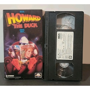 Könnte beinhalten: Eine VHS-Kassette des Films "Howard the Duck". Das Cover zeigt den Titel in Gelb und eine Illustration der Ente. Das schwarze Gehäuse zeigt auch das MCA Home Video-Logo.