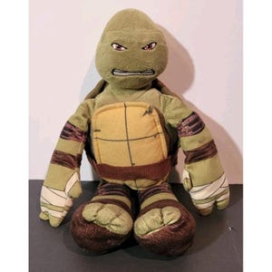 May include: A plush toy of a Teenage Mutant Ninja Turtle character. The toy is primarily green with a tan shell, brown accents, and white bandages. The turtle has an angry expression with red eyes.