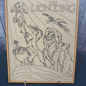 May include: A framed corkboard featuring a black line drawing of characters from "The Lion King." The artwork includes Simba, Mufasa, Timon, Pumbaa, and Zazu. The title "THE LION KING" is printed at the top.