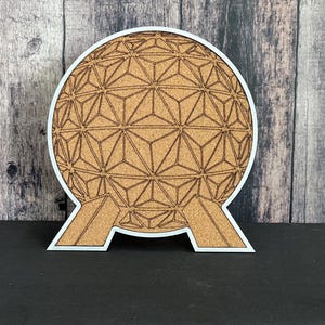 May include: A white and cork-colored wooden cutout of the Epcot Spaceship Earth attraction. The Spaceship Earth is a geodesic sphere with a geometric pattern. The cutout is on a black surface.