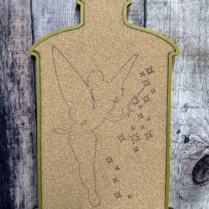 May include: A wooden decorative piece featuring a silhouette of a fairy with a wand, surrounded by stars. The fairy is inside a frame with a corkboard-like texture, outlined in green. The background is a wooden surface.