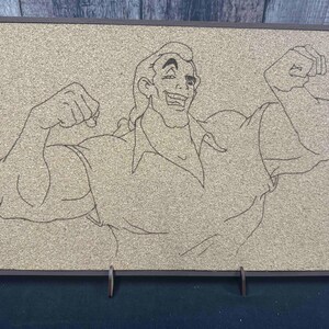 Puede incluir: Un tablero de corcho enmarcado con un dibujo lineal de un hombre musculoso flexionando sus brazos. La obra de arte est&aacute; en un marco oscuro y est&aacute; sostenida por un soporte met&aacute;lico. El fondo es de color marr&oacute;n claro.