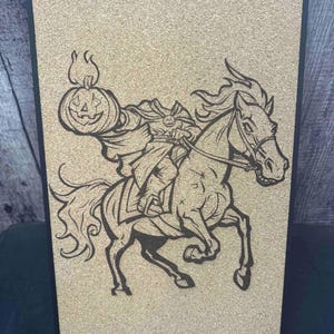 Puede incluir: Imagen en negro y marr&oacute;n del Jinete sin Cabeza sobre un tablero de corcho. La ilustraci&oacute;n representa al Jinete sin Cabeza montado a caballo, sosteniendo una linterna de calabaza. La obra est&aacute; enmarcada en negro y se mantiene en pie con un peque&ntilde;o soporte.