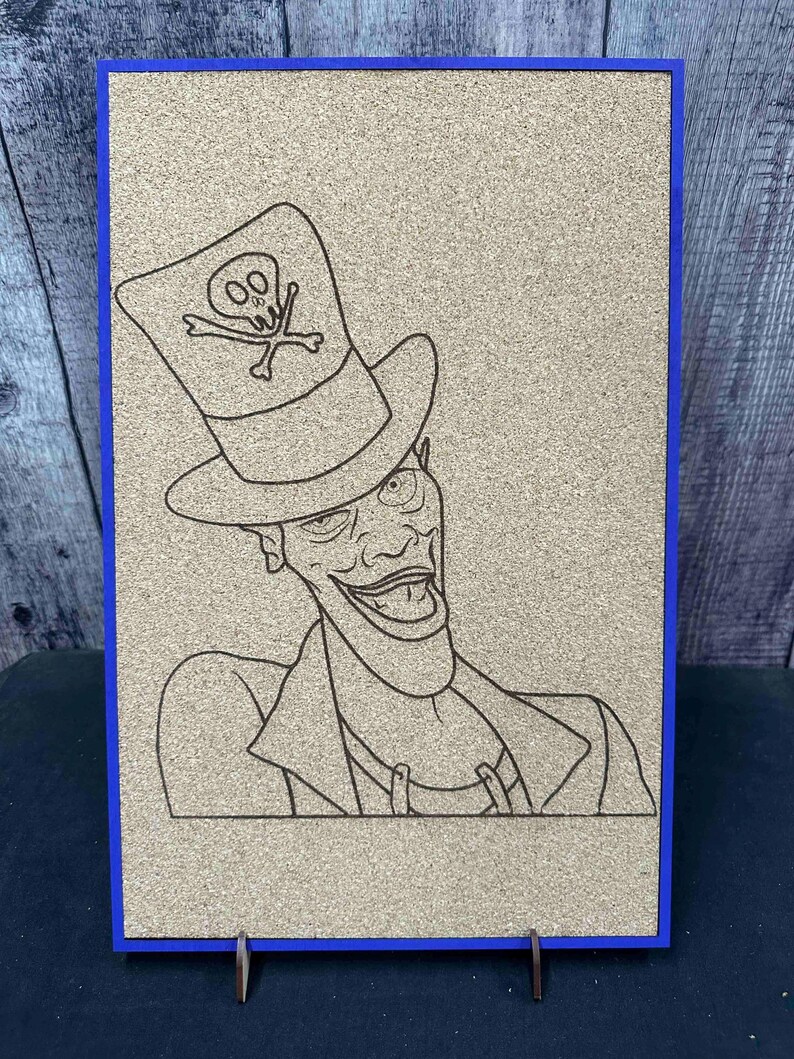 Puede incluir: Una obra de arte en tablero de corcho con marco azul presenta un dibujo lineal detallado de un personaje de dibujos animados. El personaje lleva un sombrero de copa con un emblema de calavera. La obra est&aacute; montada sobre un soporte de madera.