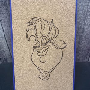 Puede incluir: Un tablero de corcho con un marco azul que presenta un dibujo lineal negro de un personaje de dibujos animados. El personaje tiene una cara grande y sonriente, con un collar. El tablero de corcho est&aacute; apoyado en un soporte.