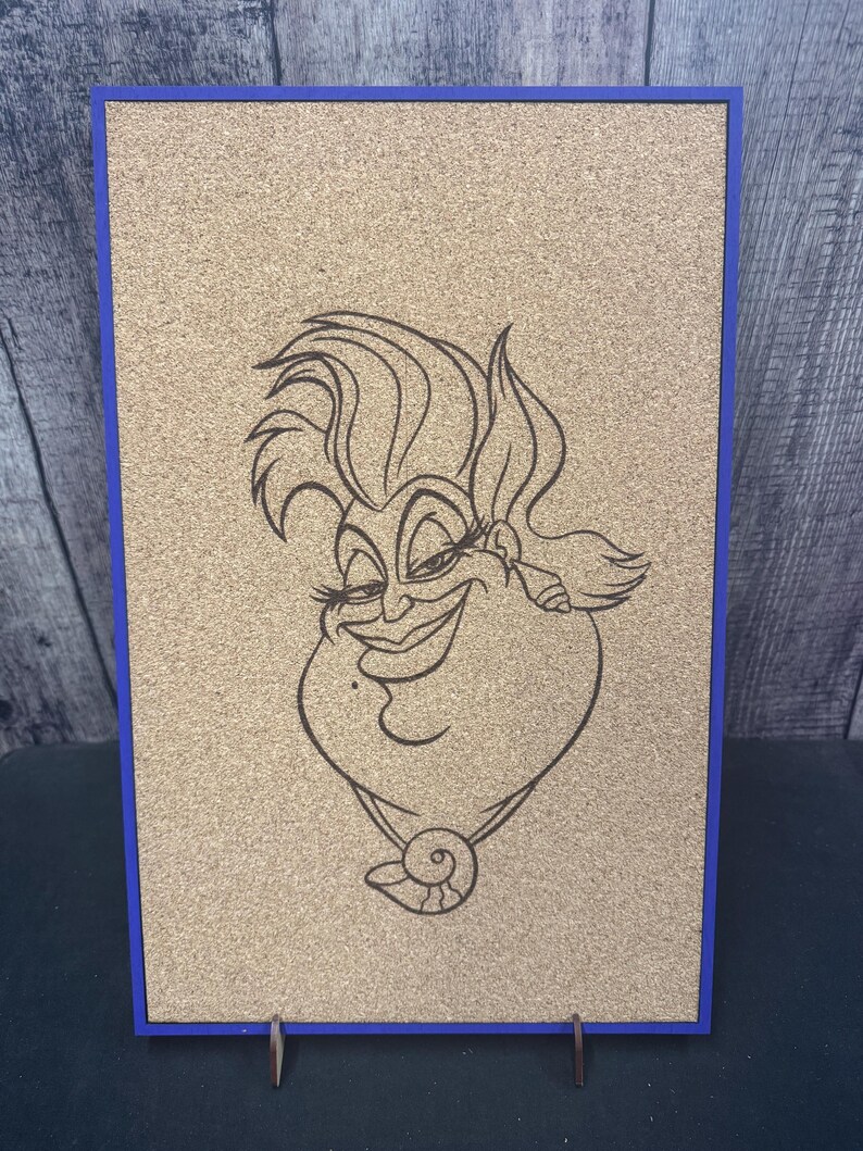 Puede incluir: Un tablero de corcho con un marco azul que presenta un dibujo lineal negro de un personaje de dibujos animados. El personaje tiene una cara grande y sonriente, con un collar. El tablero de corcho est&aacute; apoyado en un soporte.