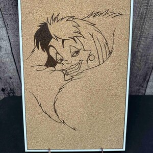 Puede incluir: Un tablero de corcho con marco blanco que presenta una imagen quemada en madera de un personaje de dibujos animados con cabello blanco y negro y una sonrisa siniestra. La obra de arte se exhibe en un soporte de madera.