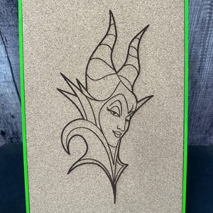 Puede incluir: Un tablero de corcho con un marco verde presenta un dibujo lineal detallado de un personaje con cuernos y una expresi&oacute;n severa. La obra de arte est&aacute; centrada en el tablero, que est&aacute; sostenido por un peque&ntilde;o soporte de madera.