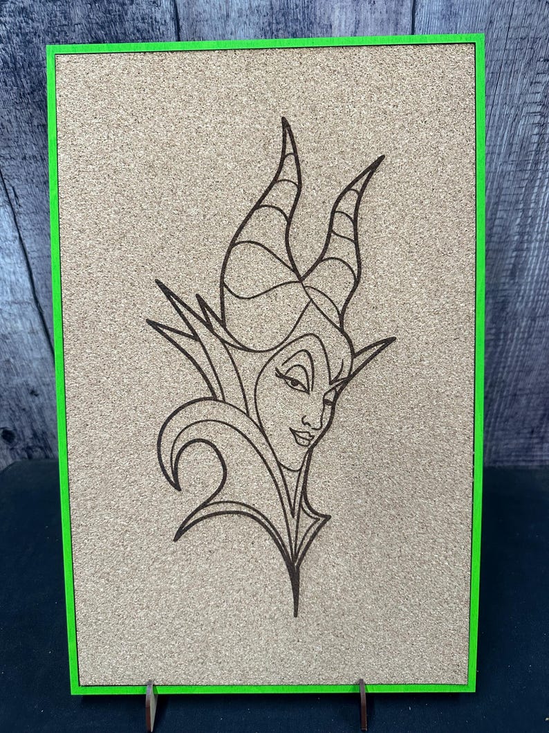 Puede incluir: Un tablero de corcho con un marco verde presenta un dibujo lineal detallado de un personaje con cuernos y una expresi&oacute;n severa. La obra de arte est&aacute; centrada en el tablero, que est&aacute; sostenido por un peque&ntilde;o soporte de madera.