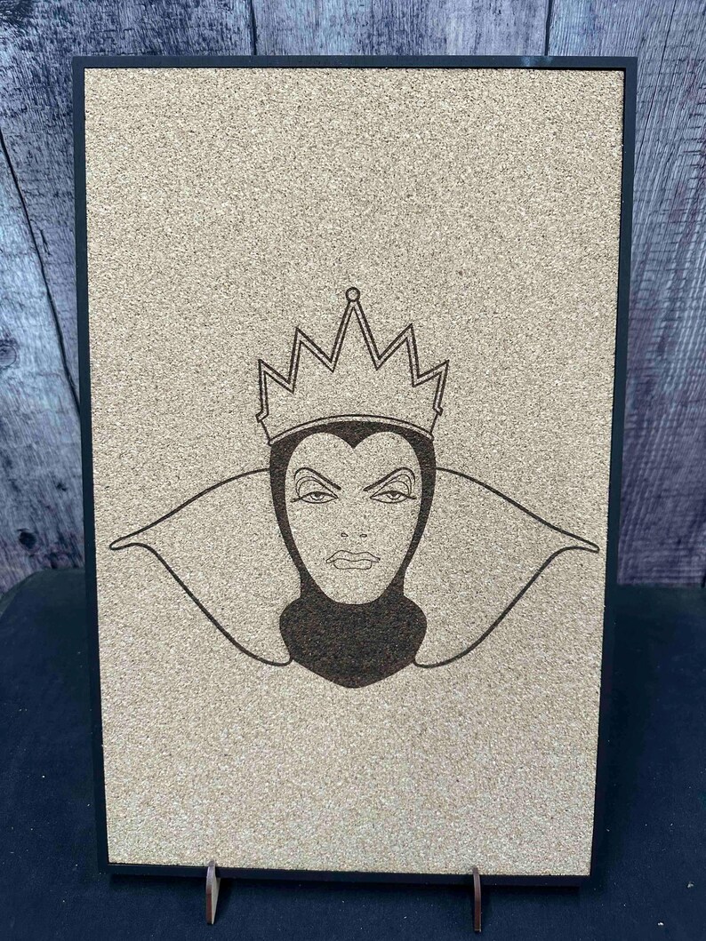 Puede incluir: Un tablero rectangular de corcho con marco negro, grabado con l&aacute;ser con la imagen de una reina severa con corona y cuello alto. La obra de arte es de color marr&oacute;n oscuro sobre corcho marr&oacute;n claro. El tablero est&aacute; sostenido por un peque&ntilde;o soporte met&aacute;lico.